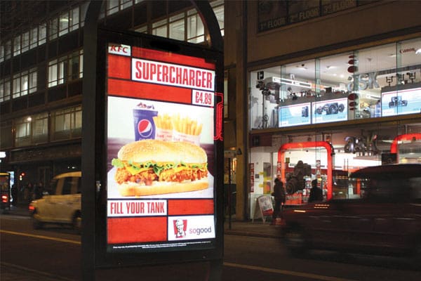 A street at night with a bus stop advertisement for KFC's Supercharger meal deal, which includes a sandwich, fries, and a Pepsi for £4.89. The ad, featuring LED backlit graphics and the slogan "Fill Your Tank," showcases the KFC and Pepsi logos. Cars and a store with illuminated signs are visible in the background.