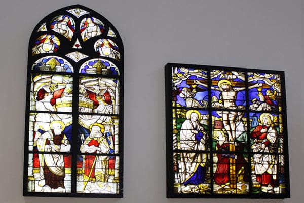 Two stained glass windows with religious scenes. The left window is tall and arched, illuminated by natural light, highlighting scenes of angels and biblical figures. The right window is rectangular, depicting various religious figures and events, set against a vibrant, colorful backdrop.