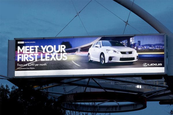 A billboard with LED backlit graphics advertises a white Lexus car under the text "MEET YOUR FIRST LEXUS" with additional text stating "From just $249 per month," accompanied by the Lexus logo. The background features a blurred cityscape at dusk, showcasing premium out of house advertising.