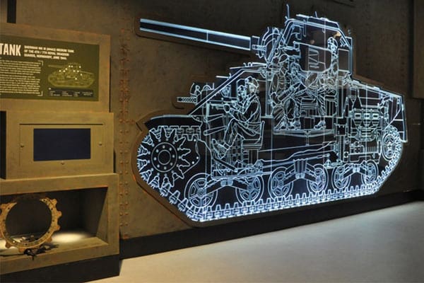 A museum display features an LED illuminated glass art piece of an intricate technical drawing of a tank. Nearby, there's an informational plaque and a display case containing a separate tank component. The environment is dimly lit, with the tank diagram prominently glowing.