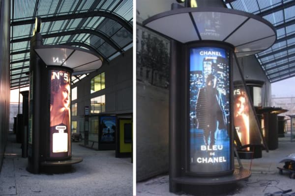 Two tall, cylindrical street advertising displays in an urban area feature illuminated LED out-of-house ads. The left display showcases a Chanel No. 5 perfume ad with a woman's face and the product bottle, while the right shows a Bleu de Chanel ad with a man in a suit at night.