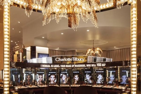 A glamorous Charlotte Tilbury makeup display in a store. The display features illuminated mirrors and LED light panels, gold accents, and rows of makeup products. Overhead, crystal chandeliers add to the opulence. Slogans and images of various makeup looks flank the products.