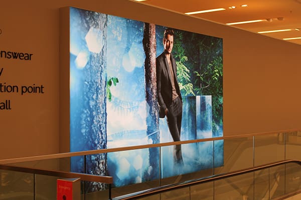 An out-of-home illuminated advertisement in a shopping mall features a man in a suit, standing confidently against a scenic, misty forest backdrop. The LED backlit graphics ad is prominently displayed above an escalator, with minimal text on the left side of the board.