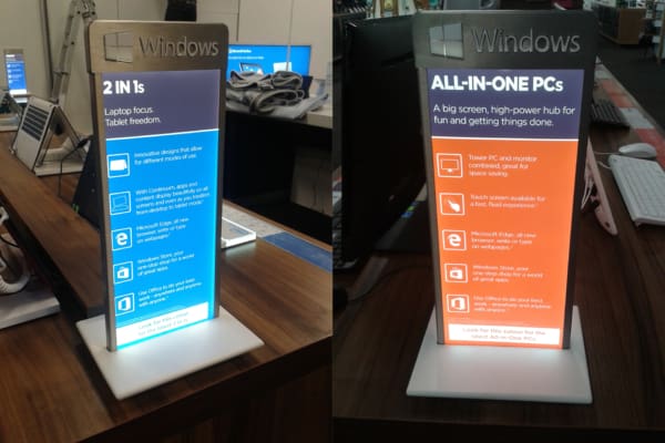 A photo of two illuminated display stands with LED backlit graphics for Windows computers in a store. The left stand promotes "2 IN 1s," highlighting features like versatile design, touch screen, and internet browsing. The right stand advertises "ALL-IN-ONE PCs," emphasizing large screen and connectivity.