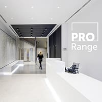 A modern, minimalist office lobby with white walls and floors, and a black ceiling section with recessed lighting. A person with long hair walks towards the reception desk. The text "PRO Range" is visible on the right side of the image.