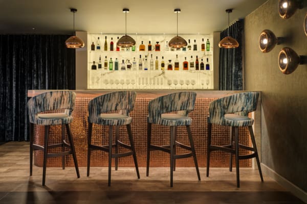 A stylish bar featuring an illuminated backlit marble shelf displays a variety of liquor bottles and glassware. Modern pendant lights hang from the ceiling, adding a sophisticated touch.