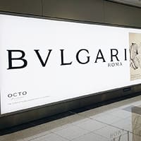 A large backlit billboard advertisement for Bulgari Roma featuring bold black text and a partial image of a watch on the right side. The ad is situated in what appears to be an indoor public space with a tiled floor.