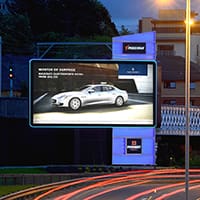 An illuminated outdoor billboard displays an advertisement for a silver luxury car against a dark background with white text and branding. The billboard is situated along a busy, curved road with streetlights, nearby buildings, and greenery in the background.
