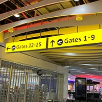 A brightly lit airport terminal sign, featuring a yellow background with black text and airplane icons for each gate group, displays directions to Gates 1-9 to the right and Gates 22-25 to the left. The sign uses an LED light sheet for illumination. The ceiling boasts curving architectural details and various lights.