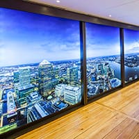 A bright, modern room with wooden flooring and large windows offers a panoramic photo of a city skyline at dusk illuminated by LED lighting.