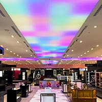 A brightly lit modern retail store interior with various display counters and shelves. The ceiling features vibrant, multicolored lighting, casting a pink and purple hue throughout the space. The store appears spacious and well-organized.