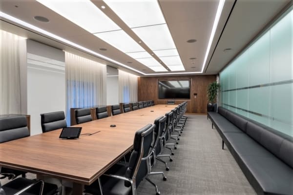 The boardroom at Carlyle Asset Management in London is a modern conference room with a long wooden table surrounded by black leather chairs. Featuring a large flat-screen TV on a wooden wall, floor-to-ceiling frosted glass panels on one side, and a bench along the opposite wall, the space is brightly lit by custom-built modular framed systems from Bright Green Technology.