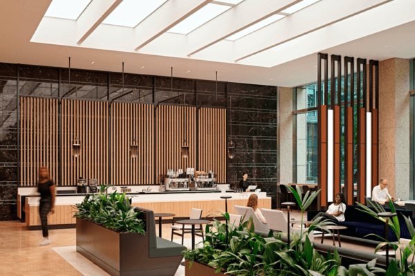 A modern, spacious office lobby features a sleek coffee bar with wooden slats on the wall, contemporary furniture, and indoor plants. People are sitting and enjoying the space, which is well-lit with natural light from large windows and skylights enhanced by tuneable white LED Light boxes.