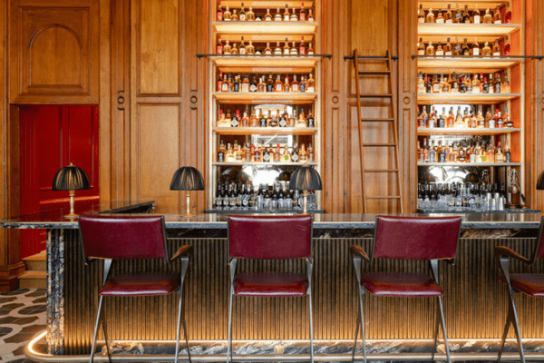 A stylish bar features a wooden back wall with shelves filled with bottles of liquor, illuminated by LED shelf-lighting. The front counter is marble with ribbed paneling, flanked by three red upholstered bar stools. The floor has a black and white geometric pattern.