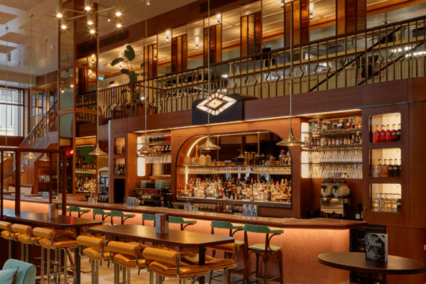 A stylish bar interior featuring wooden elements, tall stools at a sleek counter, and plush seating with colorful cushions is beautifully complemented by PRO Range LED Light Panels backlighting Dishoom’s Permit Bar. Large windows and a mezzanine level enhance the spacious atmosphere.