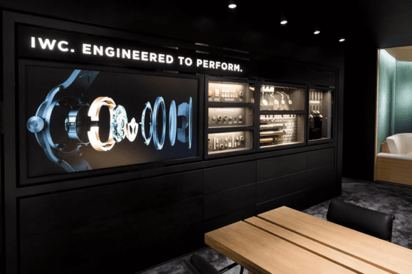 A modern showroom interior featuring a wall display with "IWC. Engineered to Perform." showcases mechanical watch components, backlit by four custom slimline lightboxes. The contemporary space includes a seating area and wooden table set, highlighting prime features with elegant lighting.