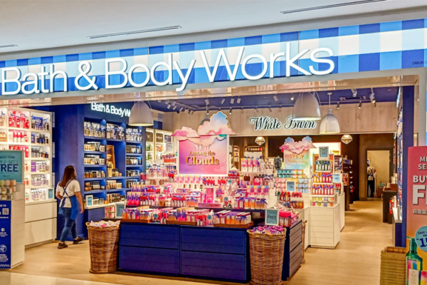 A Bath & Body Works store with a bright, LED backlit entrance and a blue and white checkered exterior. The storefront displays various colorful products, promotional signs for "Buy 2, Get 1 Free" and "150 Mix & Match." A person is browsing on the left.