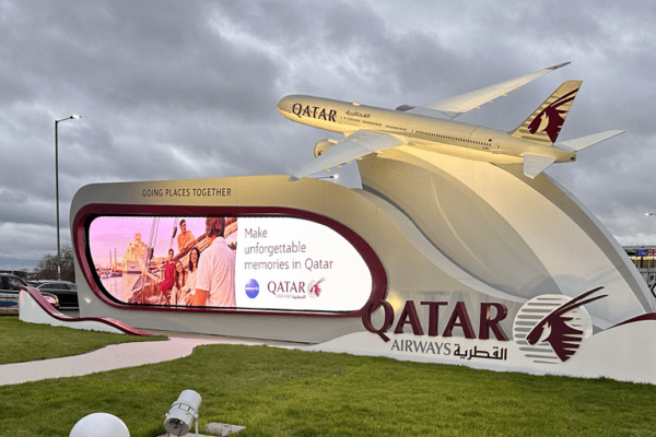 A large outdoor display showcases a 9-meter-long scaled model of a Boeing 777-9 aircraft perched atop a curved structure, above a 12-meter LED screen. It highlights Qatar Airways' message about making unforgettable memories in Qatar. The cloudy sky looms overhead as several cars are parked nearby.