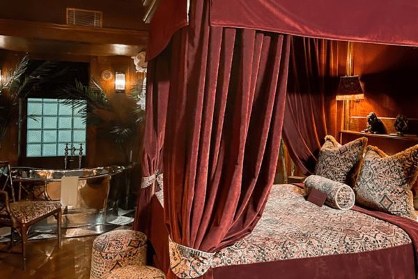 A luxurious, dimly lit bedroom at the House of Gods Hotel features a large canopied bed adorned with patterned pillows and a rich, dark comforter. Dynamic white LED light panels with DMX drivers adjust to replicate the time of day, illuminating a freestanding bathtub by window-side curtains.
