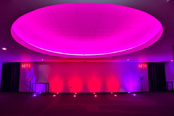 A modern room glows with energy-efficient, enhanced uplighting from a large oval ceiling light in pink and purple hues. Two small digital displays titled "NFT1" adorn opposite walls, crafting a vibrant, futuristic atmosphere.