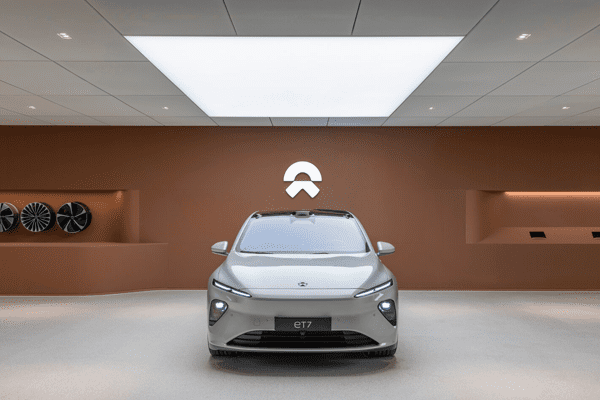 A sleek gray electric car, the NIO ET7, is showcased in a modern showroom with backlit surfaces and a minimalist design. A large ceiling light illuminates the space, highlighting the brown backdrop with the NIO logo and wheels mounted on the left wall.