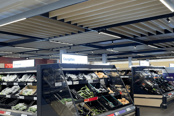 A grocery store produce section with shelves filled with various vegetables, including zucchinis and squash. The ceiling features striped patterns and bright lighting. Signs label different sections, such as "Courgettes.